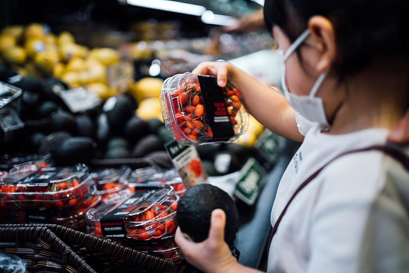 可爱的小亚洲女孩戴着保护面罩在超市购物。她正在农产品货架上采摘新鲜的有机牛油果和一盒枸杞浆果图片素材