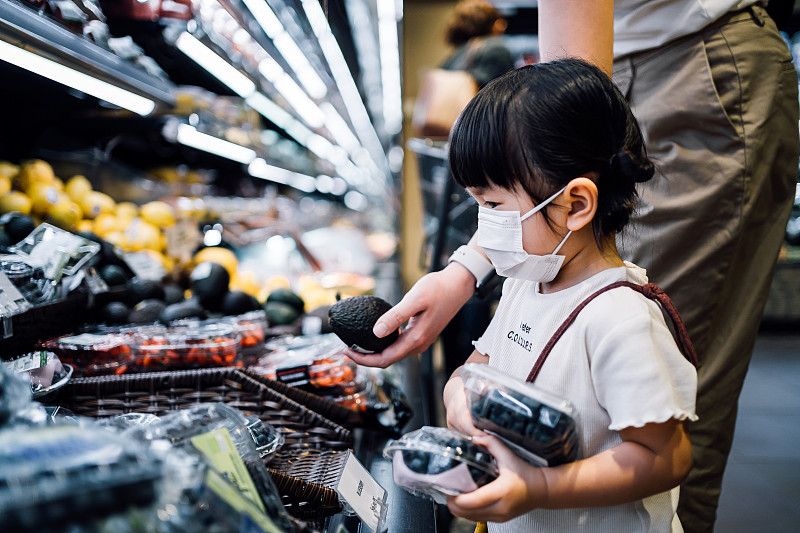 Cute little Asian girl with protective face mask grocery shopping with her mother in a supermarket. They are choosing fresh organic blueberries and avocados in the produce aisle图片素材