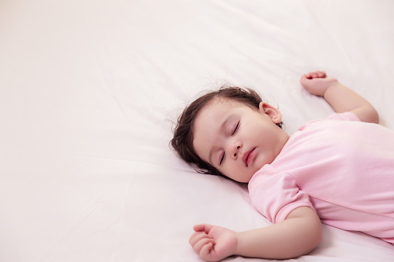 可爱天真的小女孩睡在床上，做着好梦。可爱的混血婴儿宝宝休息舒适，轻松安宁。可爱的蹒跚学步的小女孩穿着粉红色的婴儿连衣裙。本空间图片下载