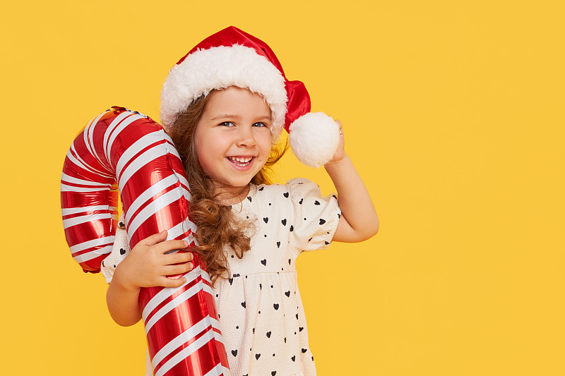 一个可爱的小女孩穿着花哨的衣服，戴着圣诞老人的帽子，拿着一个黄色背景上的充气拐杖糖。2021年新年假期概念和儿童新年礼物广告图片下载