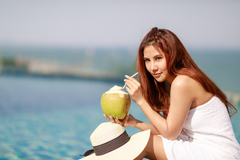 年轻的亚洲女子放松在游泳池与椰子汁。图片下载