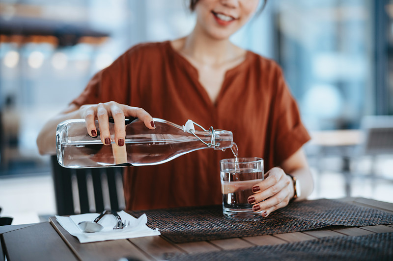 在户外餐厅，年轻的亚洲女子将瓶子里的水倒进玻璃杯里。健康的生活方式，保持水分图片下载