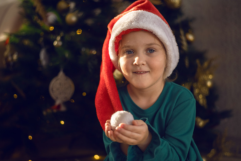 一个戴着圣诞老人帽子的小女孩在圣诞树下许愿。图片下载