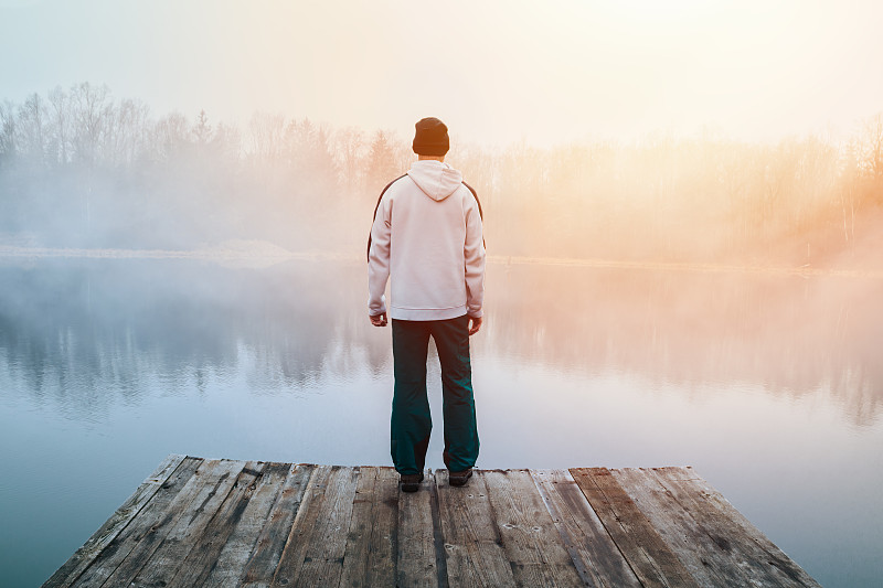 年轻男子在帽衫，帽子和裤子站在木码头在池塘岸边与忧郁的雾日出。捷克晨景图片下载