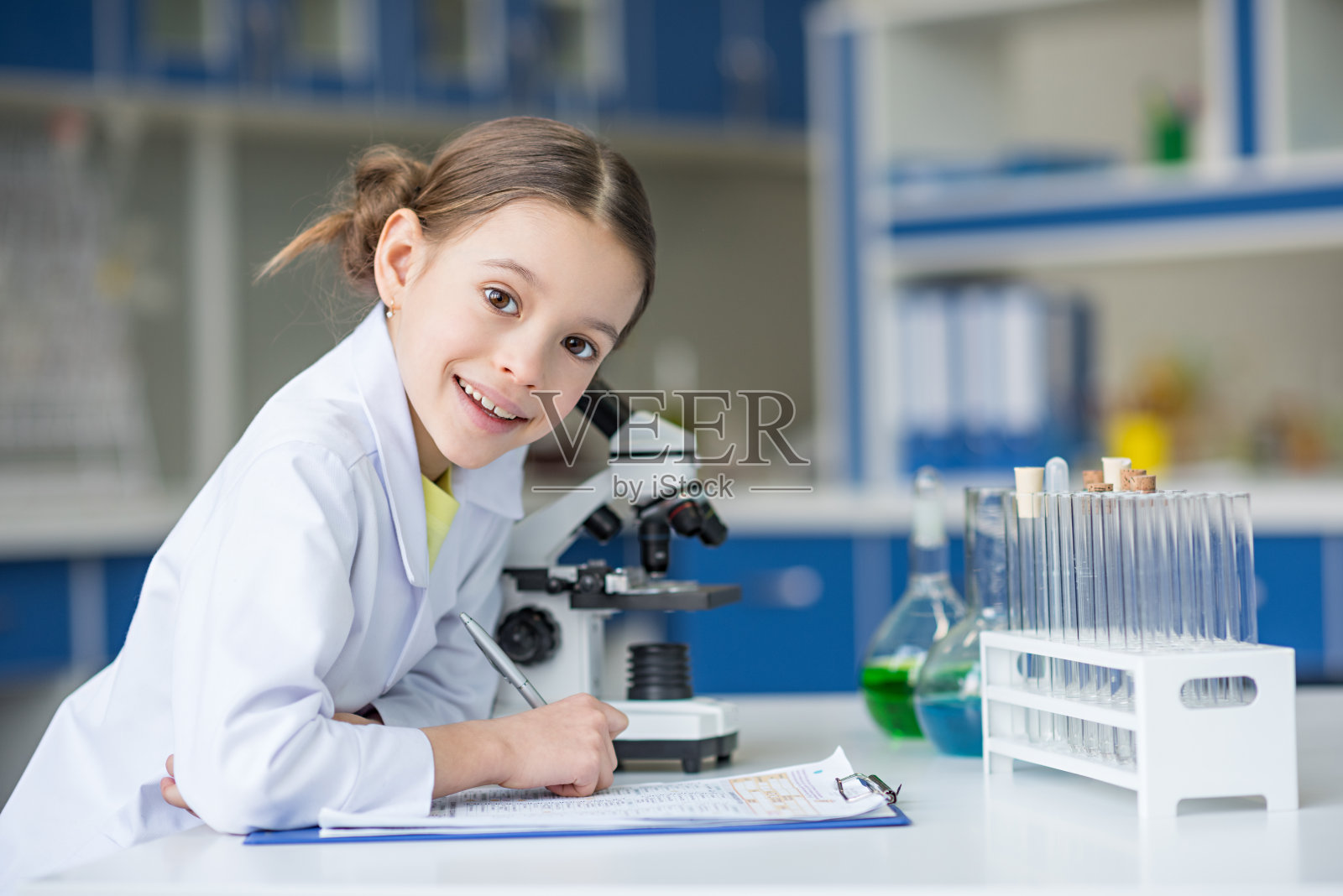 穿着实验服的女科学家在写字板上写字，对着镜头微笑照片摄影图片
