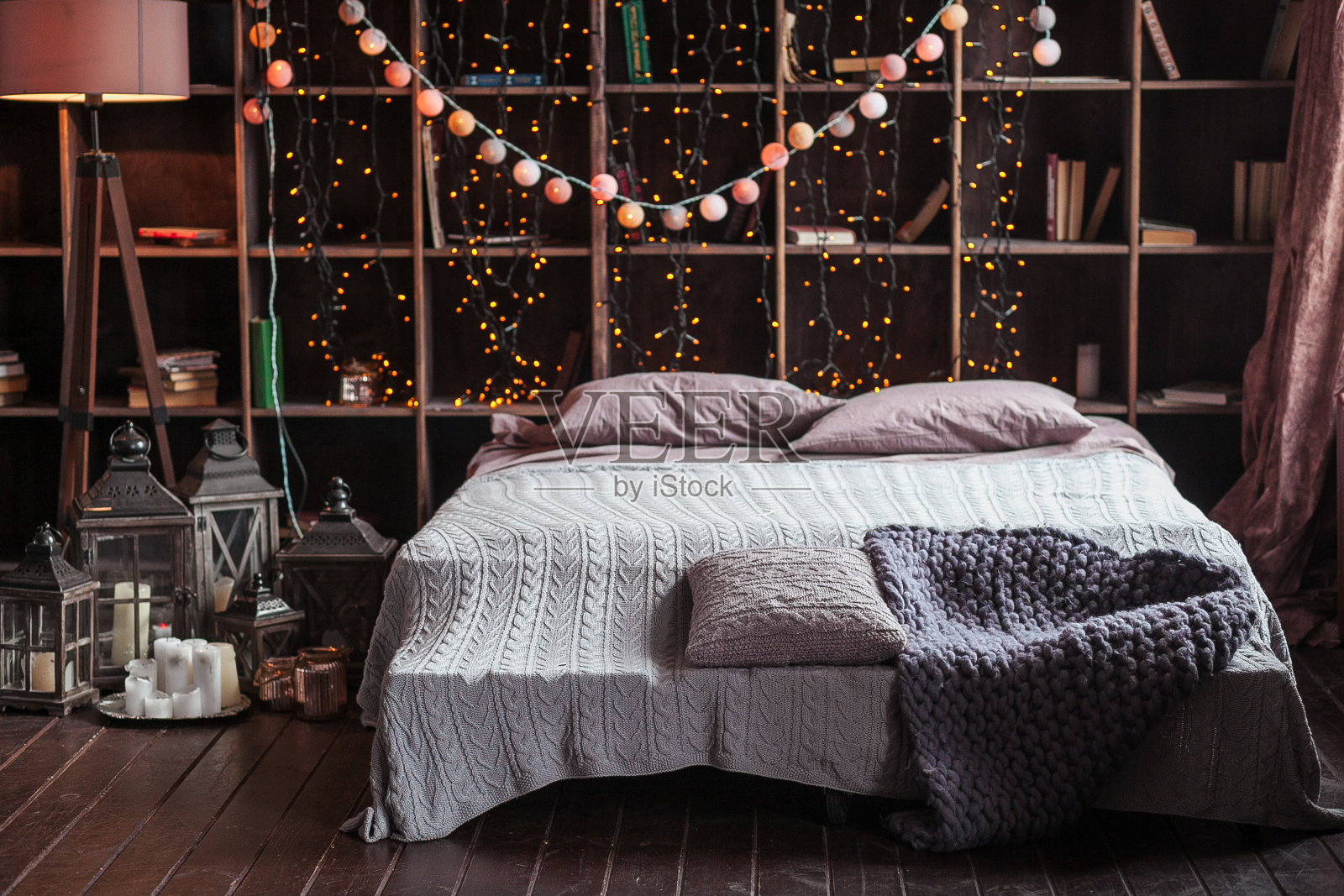 舒适，舒适，室内和假日的概念-舒适的卧室，床和花环灯在家里。床后面放书的架子。床边有蜡烛、一盏灯和一个灯座。格子手工编织的照片摄影图片