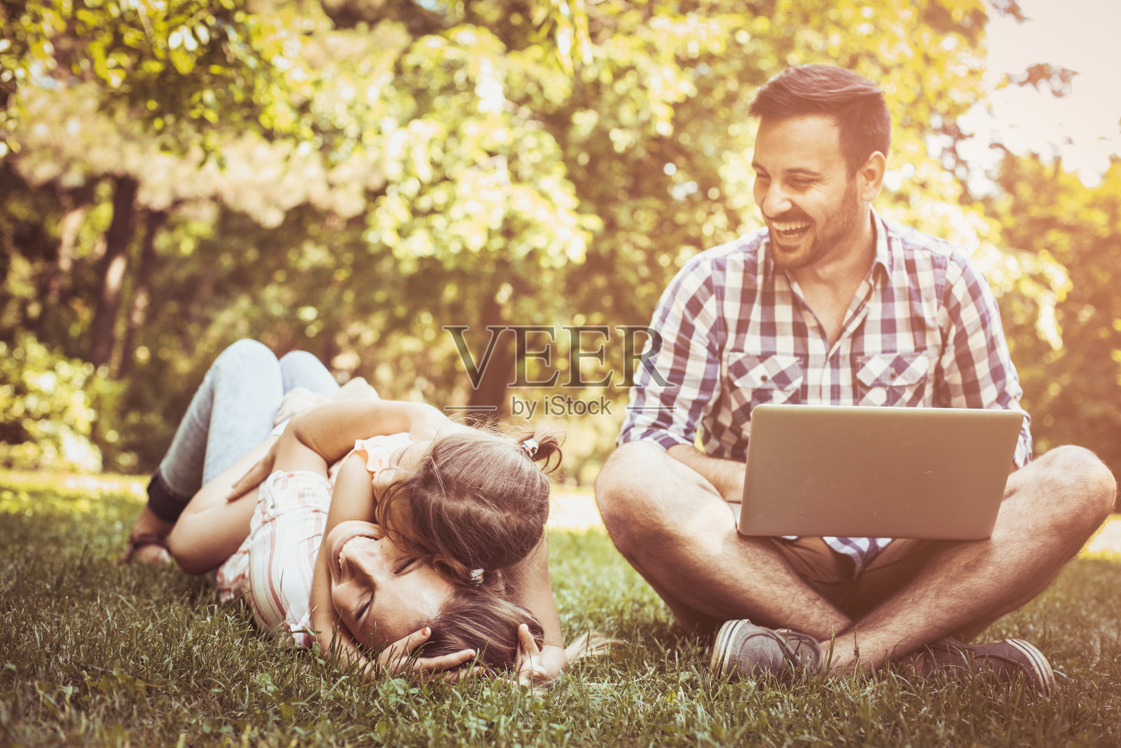 有一个孩子的幸福家庭。父亲坐在草地上用笔记本电脑。照片摄影图片