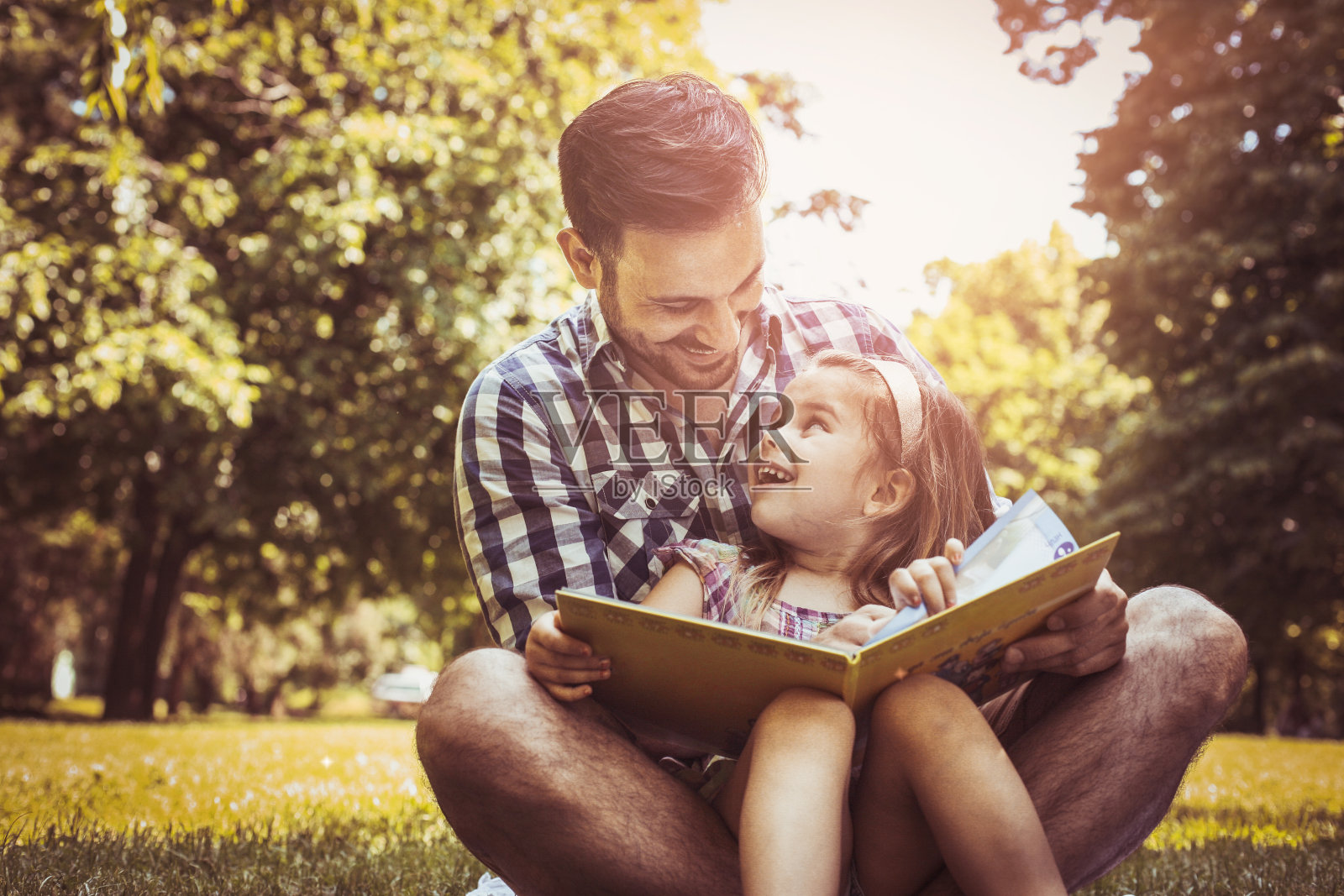 单身父亲和小女儿坐在草地上读故事书。小女孩坐在爸爸腿上。照片摄影图片