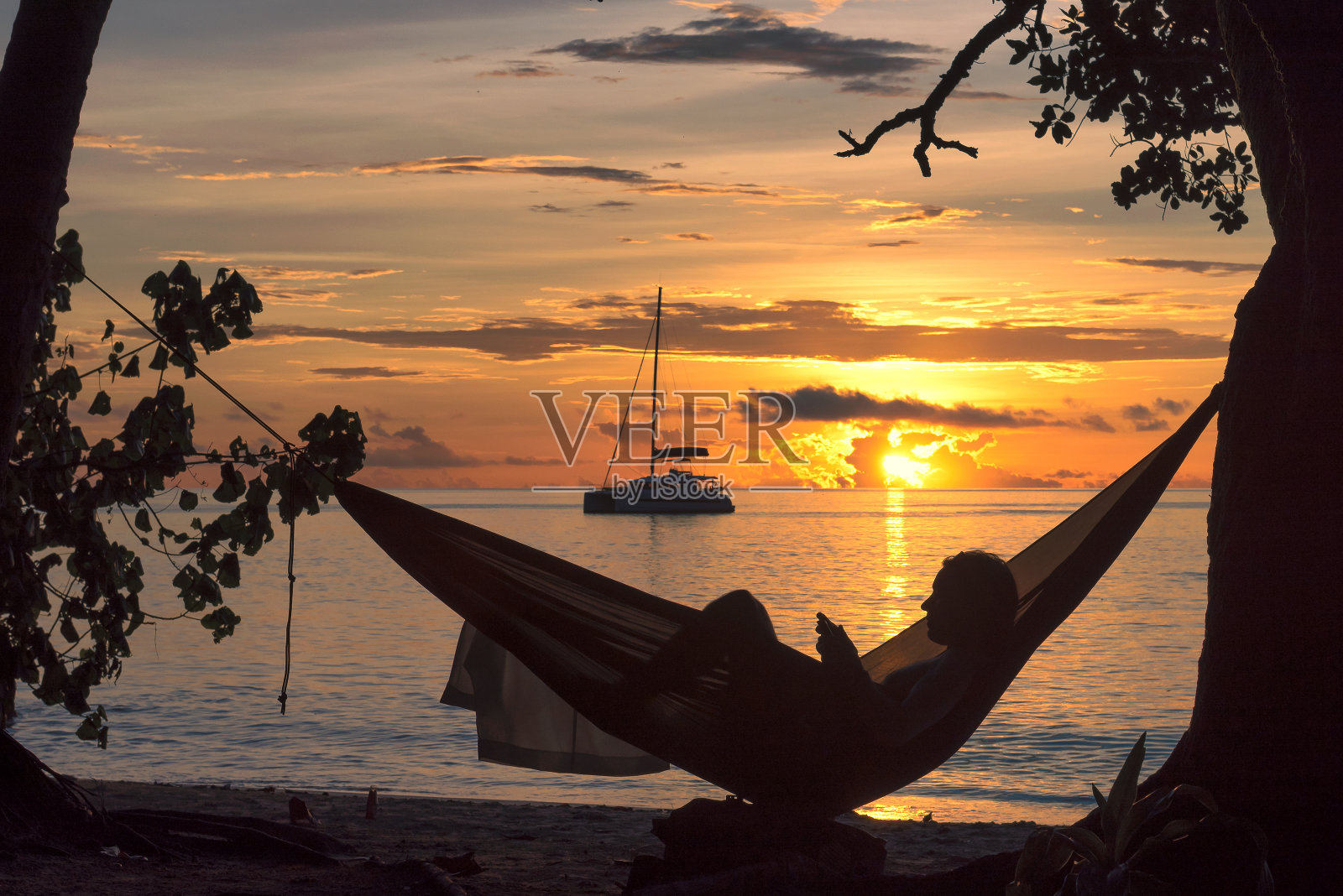 海滩度假，一个人的剪影在吊床上阅读日落在热带岛屿。照片摄影图片