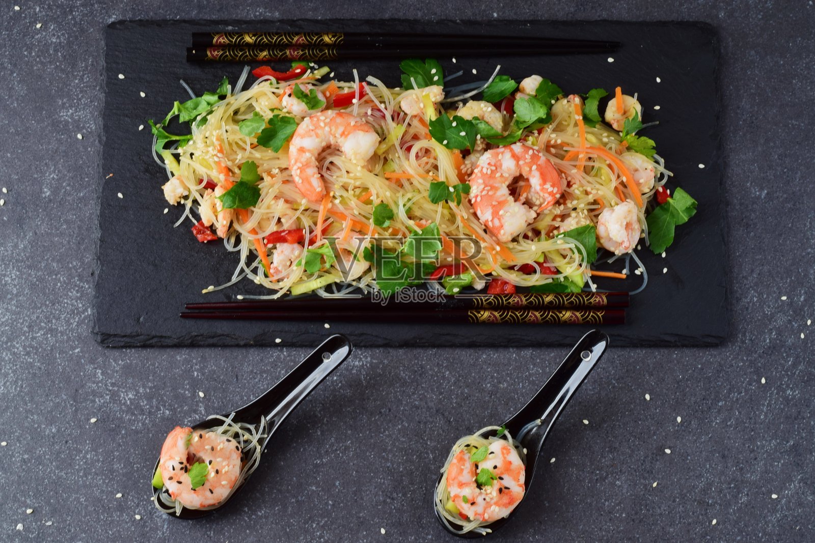 在灰色抽象背景上，用传统汤勺盛着黑色石盘上的虾仁和蔬菜面条。健康食品的概念照片摄影图片