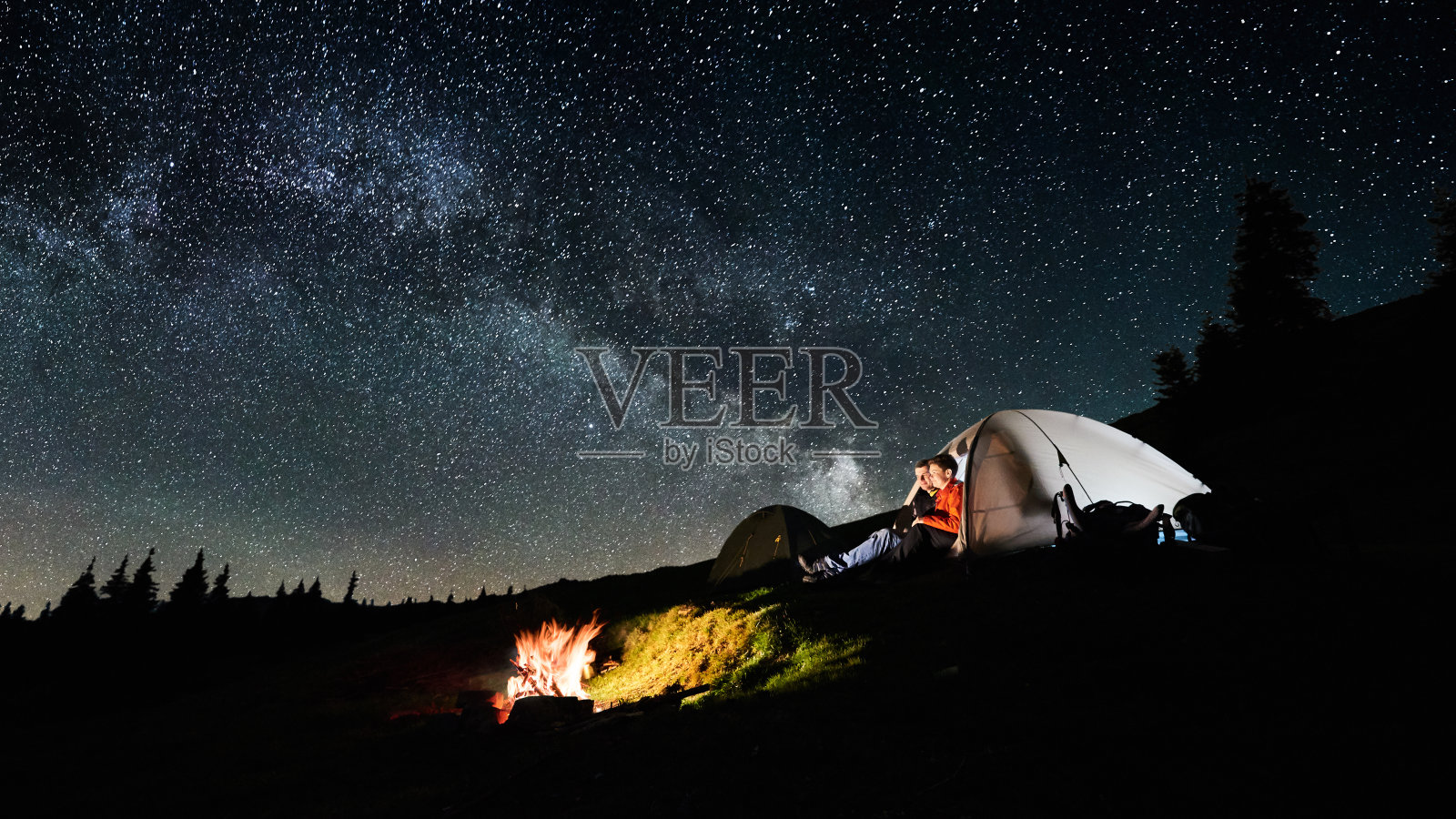 晚上露营。男男女女游客坐在篝火旁的灯火通明的帐篷里，在满是星星和银河的神奇夜空下。长时间的曝光。图片宽高比16:9照片摄影图片