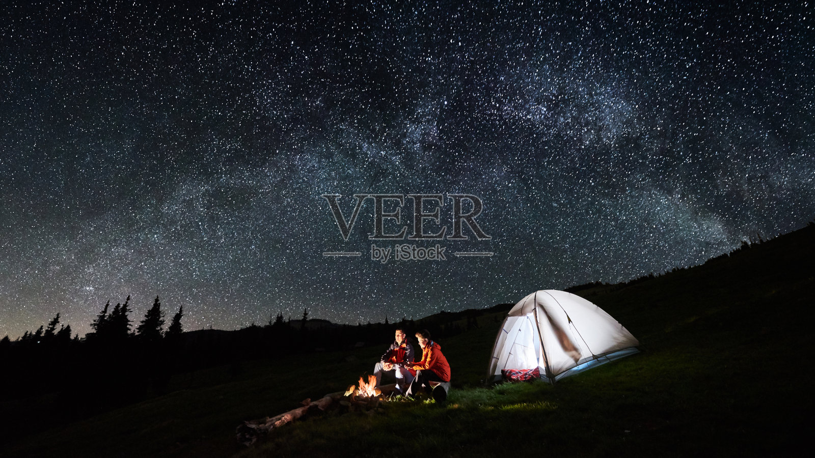 晚上在山里露营。在满是星星和银河的美丽夜空下，游客夫妇在明亮的帐篷旁的篝火旁休息。低光。图片宽高比16:9照片摄影图片