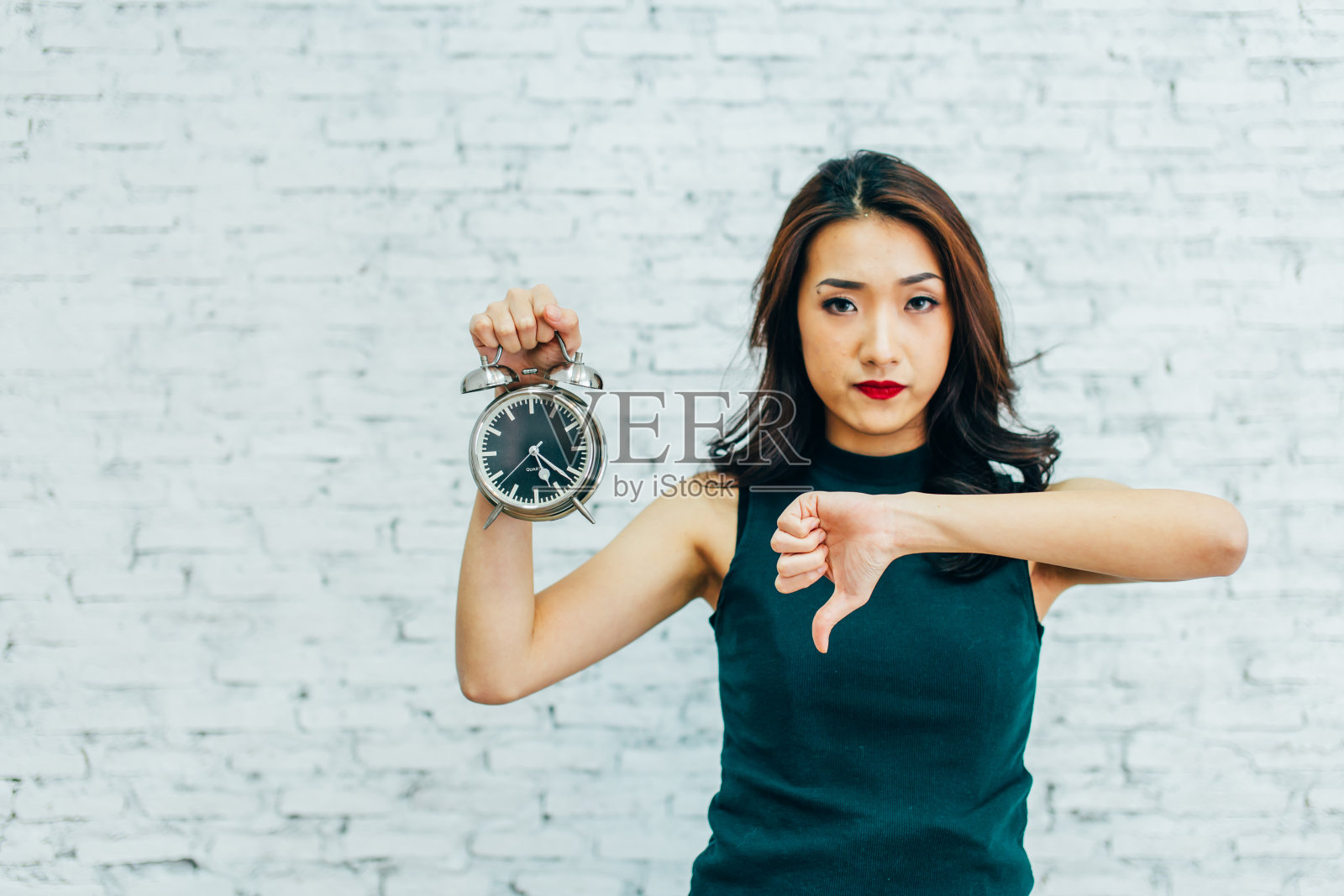 亚洲商务女性拇指朝下，手持闹钟——表示对时间管理不满意照片摄影图片