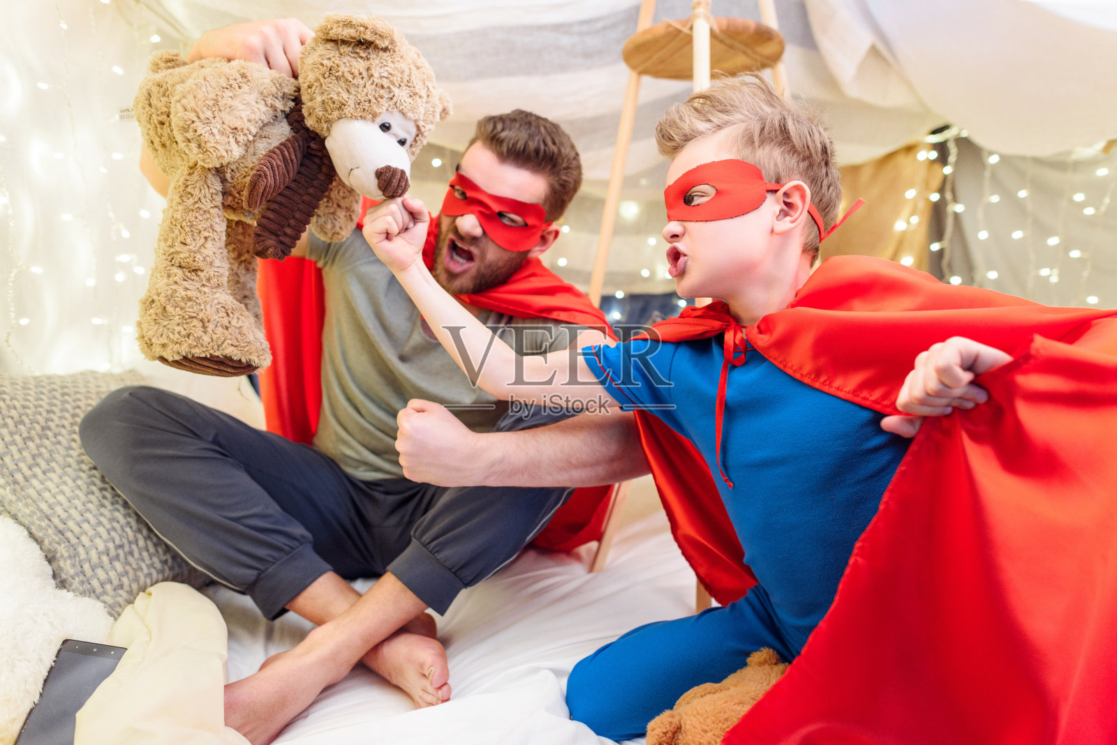 父亲和儿子穿着超级英雄的服装在毯子堡垒里玩泰迪熊照片摄影图片