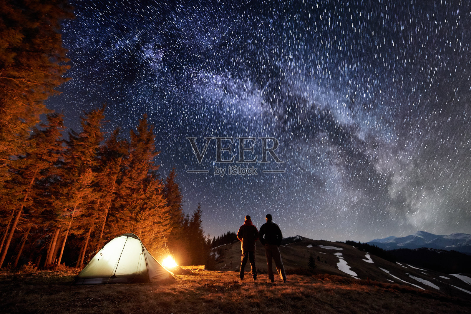 两名男性游客晚上在森林附近的露营中休息。小伙子们站在篝火和帐篷旁，在满是星星和银河的美丽夜空下，欣赏夜景。长时间曝光照片摄影图片