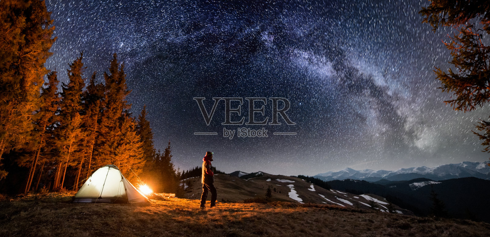 男游客晚上在森林附近的营地休息。一个人站在篝火和帐篷附近，在美丽的夜空下，满是星星和银河，欣赏夜景。全景景观照片摄影图片
