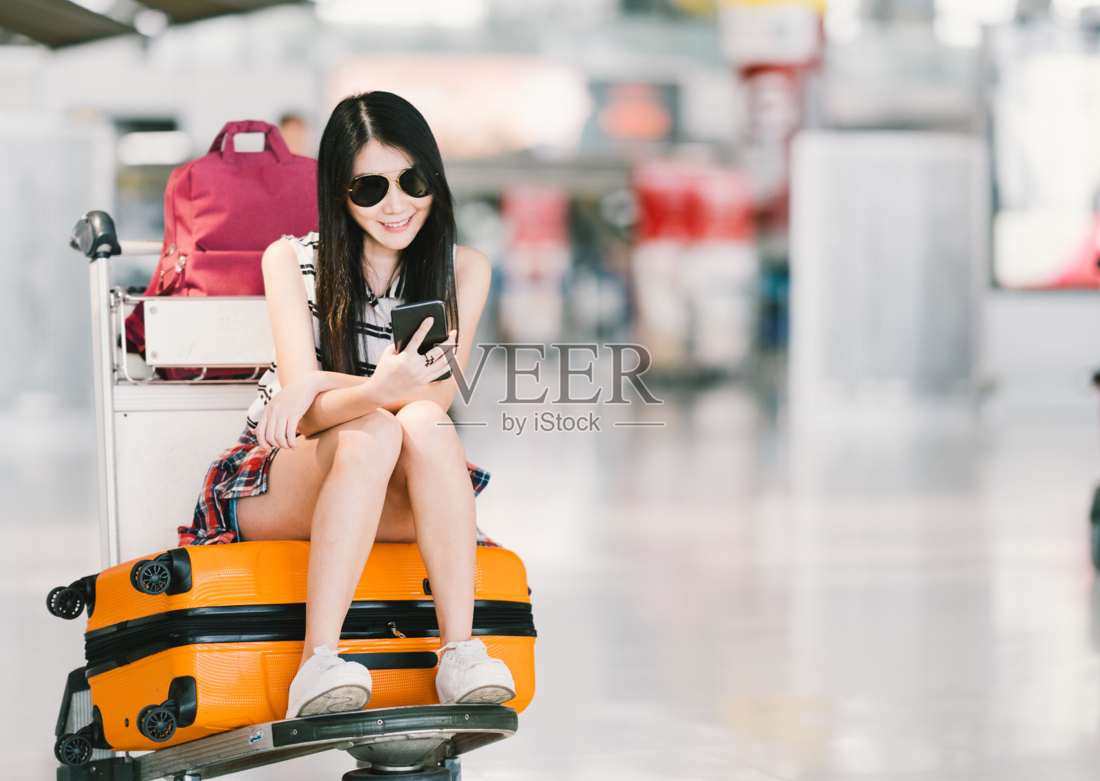 年轻的亚洲女孩使用智能手机，在机场等待航班，坐在行李手推车或行李车上。移动通信技术或出国旅游概念，具有复制空间照片摄影图片