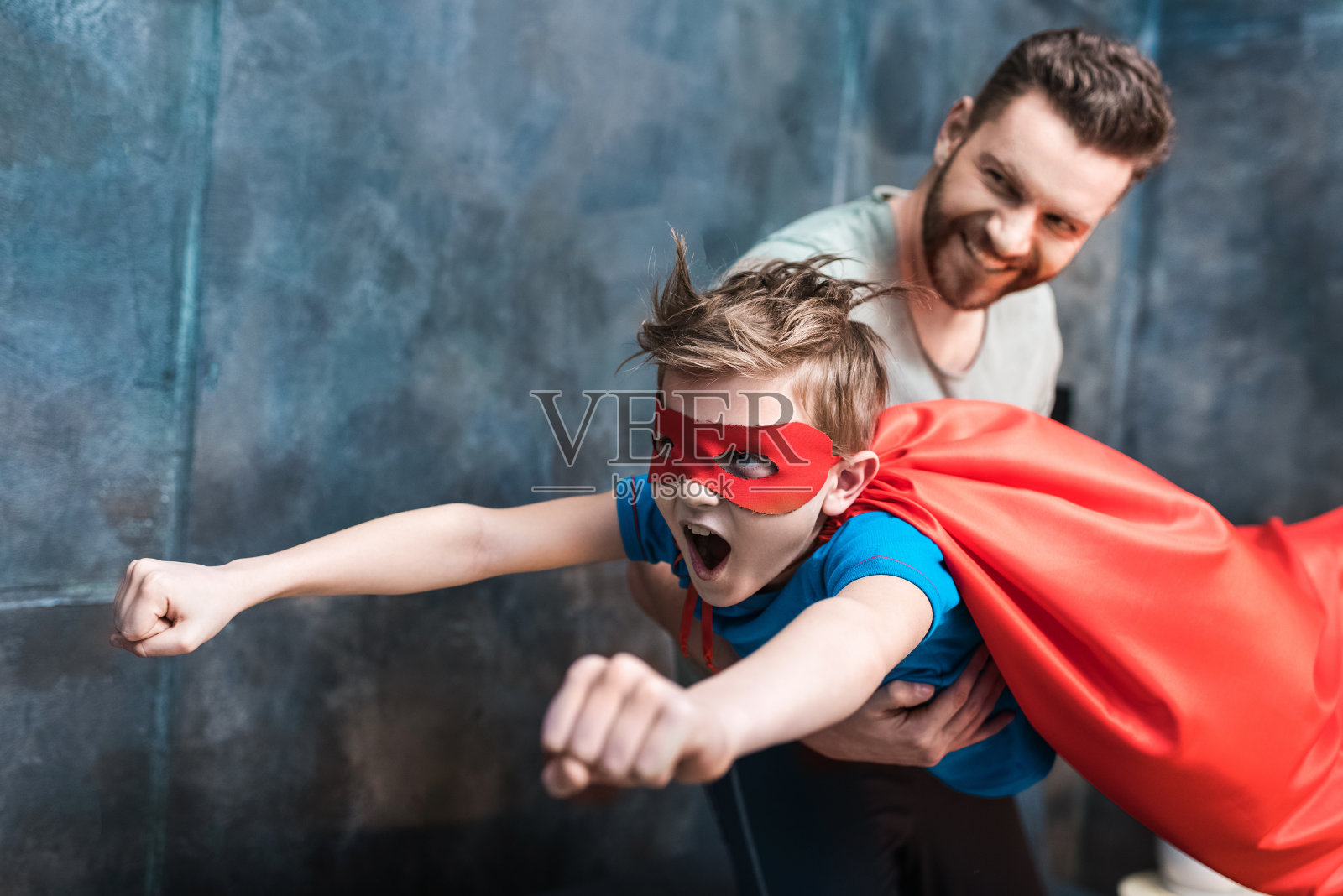 父亲抱着穿着超级英雄服装的儿子在家飞行照片摄影图片