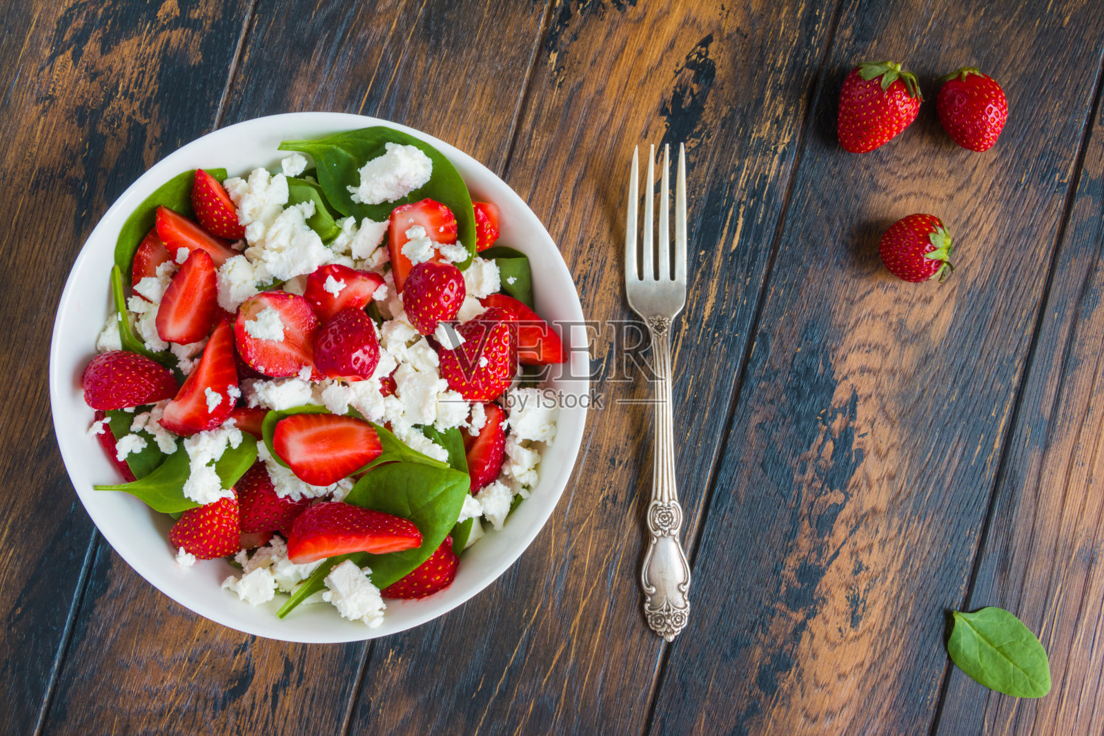 健康的素食沙拉，新鲜的草莓，绿色的菠菜叶和羊乳酪放在一个白色的碗里放在木制的乡村桌子上，俯视图。照片摄影图片
