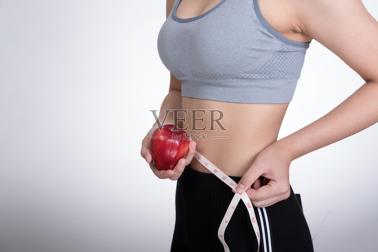 运动健身女性用卷尺量腰围。戴着红苹果的年轻女孩在白色背景下测量自己的身体。饮食、运动和健康理念照片摄影图片