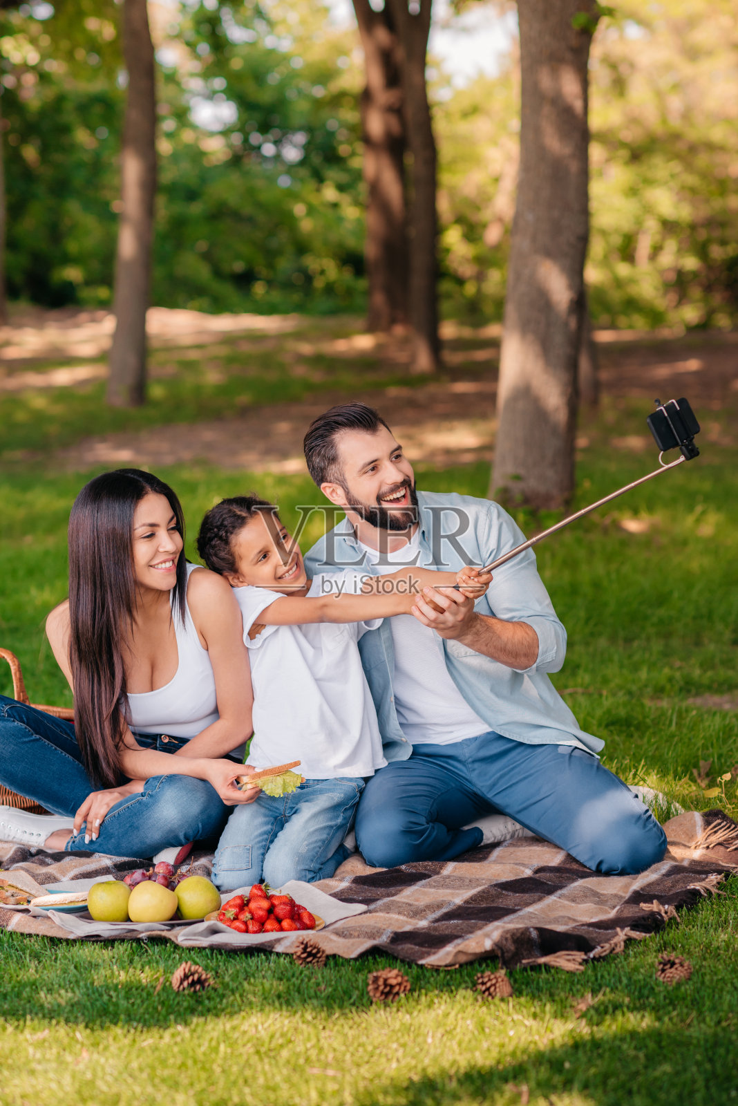 一个快乐的家庭，一个孩子在野餐时坐在格子布上用智能手机自拍照片摄影图片
