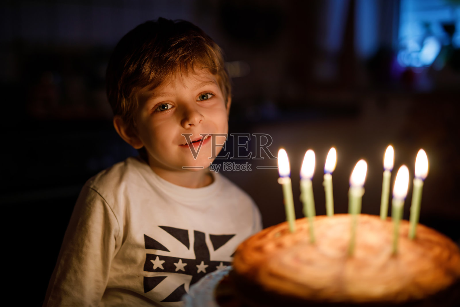 可爱的快乐的金发小男孩庆祝他的生日。孩子吹七根蜡烛在自制的烤蛋糕上，室内。照片摄影图片