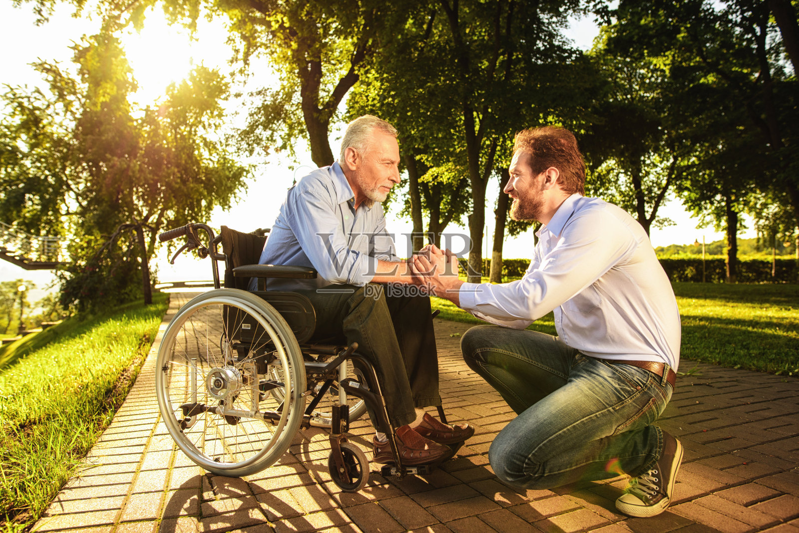 坐在轮椅上的老人和他的儿子在公园里。他们走路。一个男人牵着一位老人的手照片摄影图片