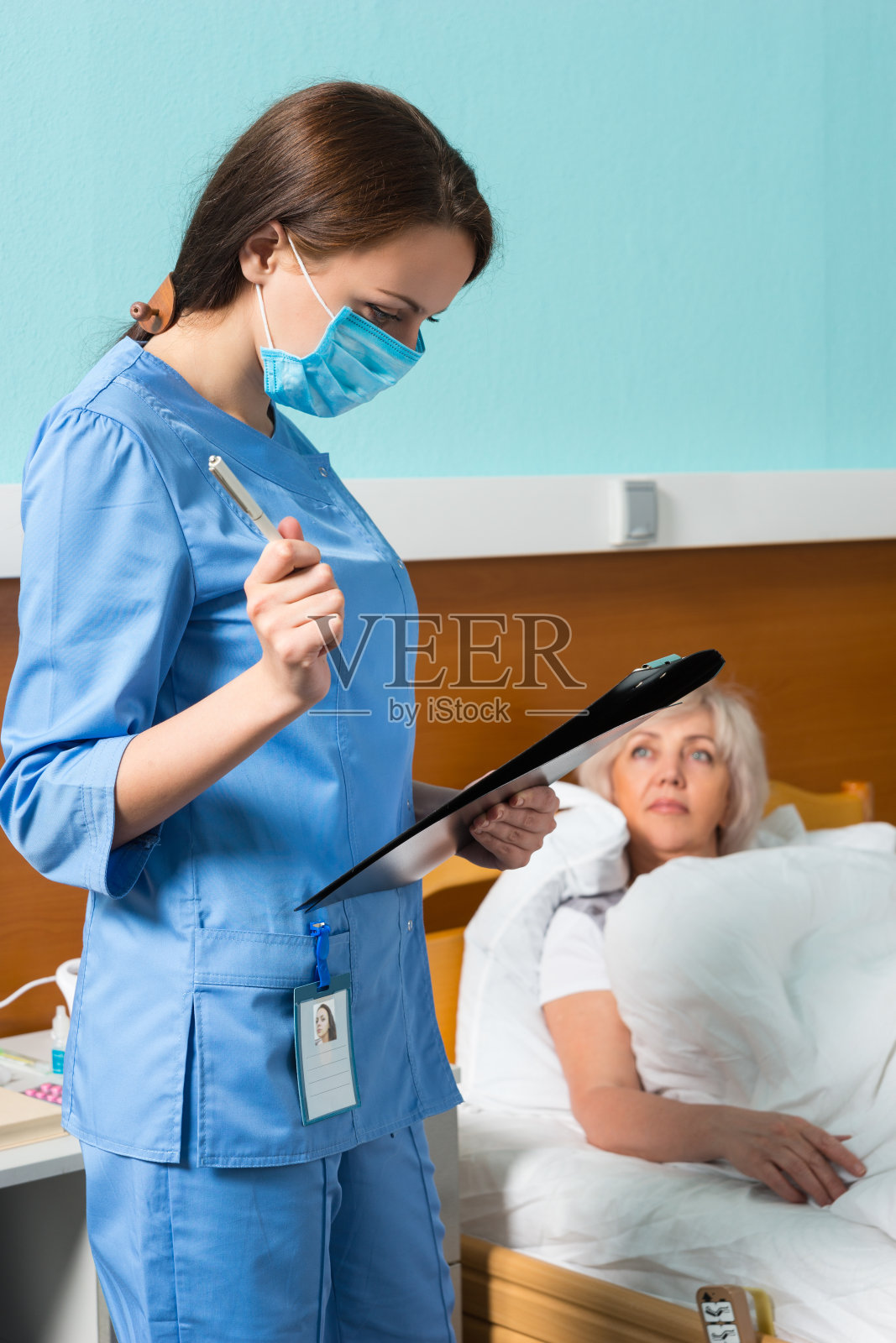 漂亮的护士穿着制服戴着口罩看剪贴板与报告，而探望她的病人照片摄影图片