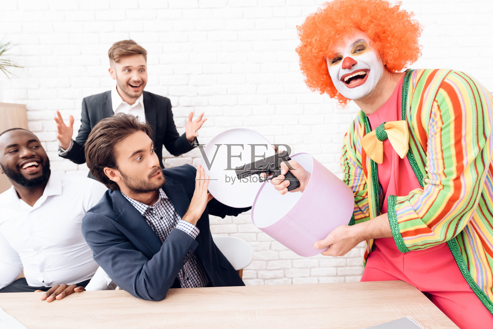 在一间明亮的办公室里，一名穿着小丑服的男子正用枪瞄准商人。照片摄影图片