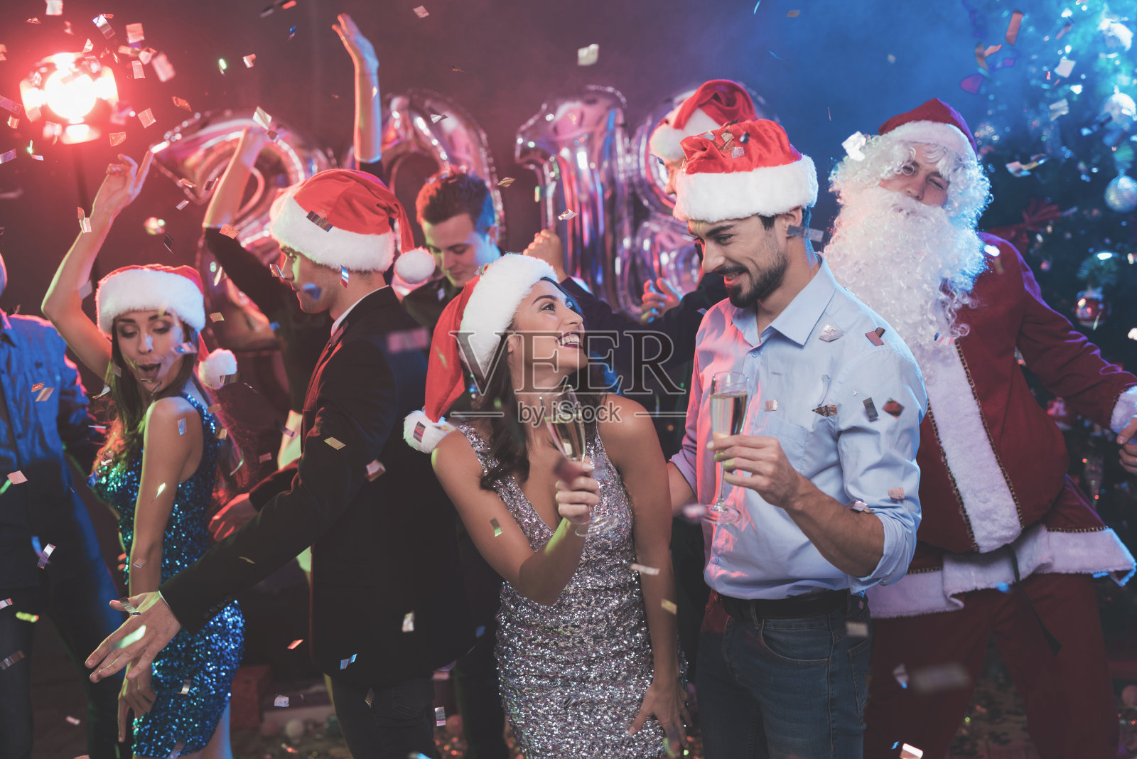 年轻的一对手拿香槟杯跳舞。在他们后面跳舞的是他们的朋友和打扮成圣诞老人的人。照片摄影图片