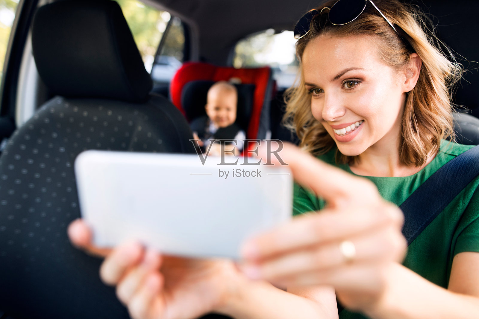 年轻妈妈拿着智能手机，车里坐着一个男婴。照片摄影图片