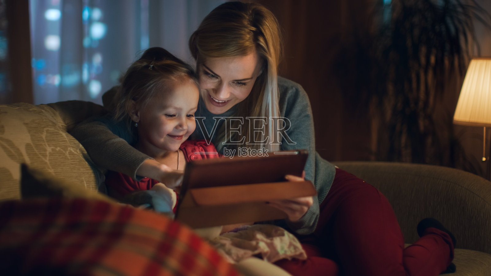 美丽的妈妈和她的小女儿正坐在客厅的沙发上，他们在用平板电脑。这是晚上，房间是舒适和温暖的。照片摄影图片