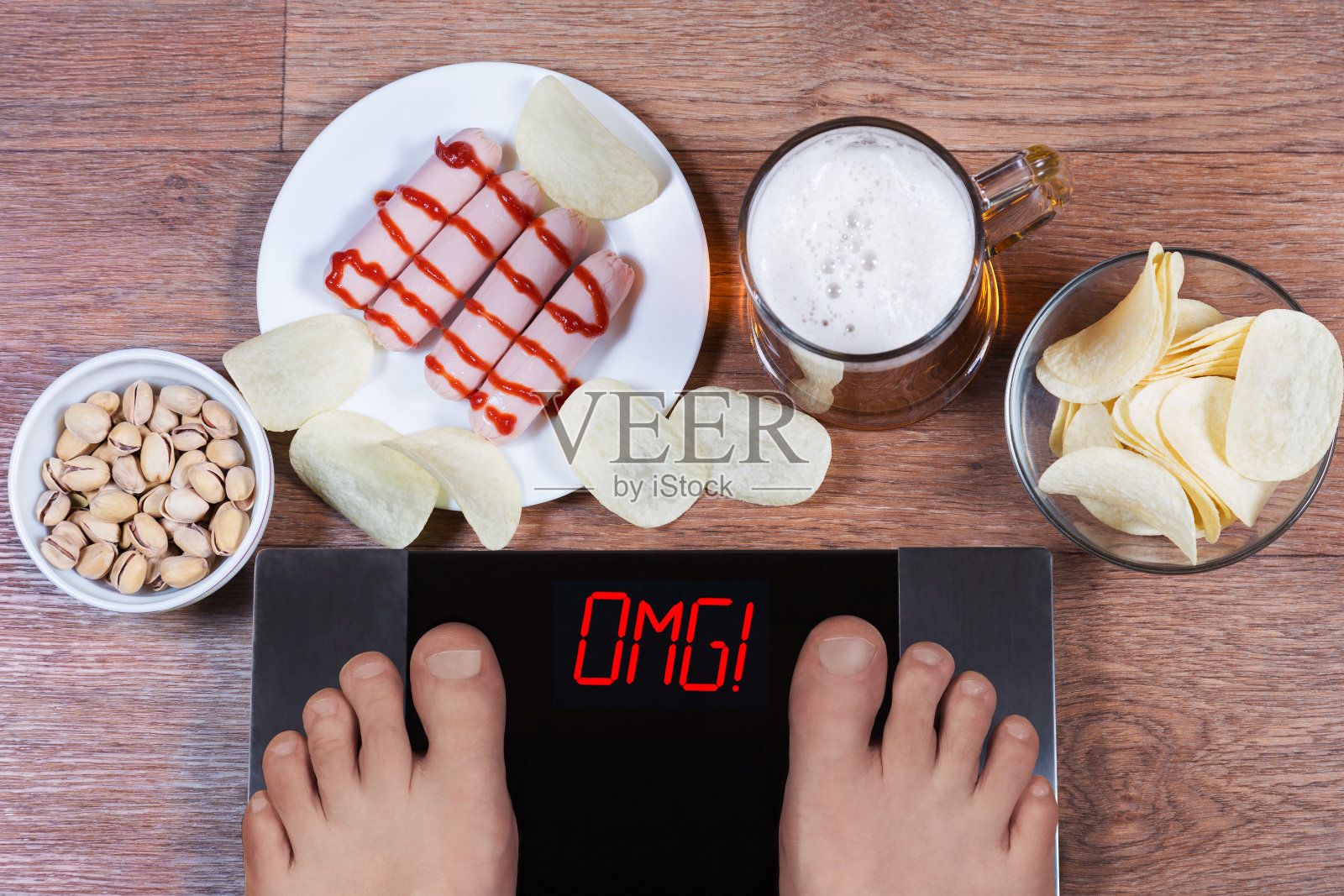 男性的脚在数字秤上，屏幕上有“omg”这个词。啤酒和装有垃圾食品(香肠、薯片、开心果)的盘子。不健康饮食和饮酒的概念。照片摄影图片