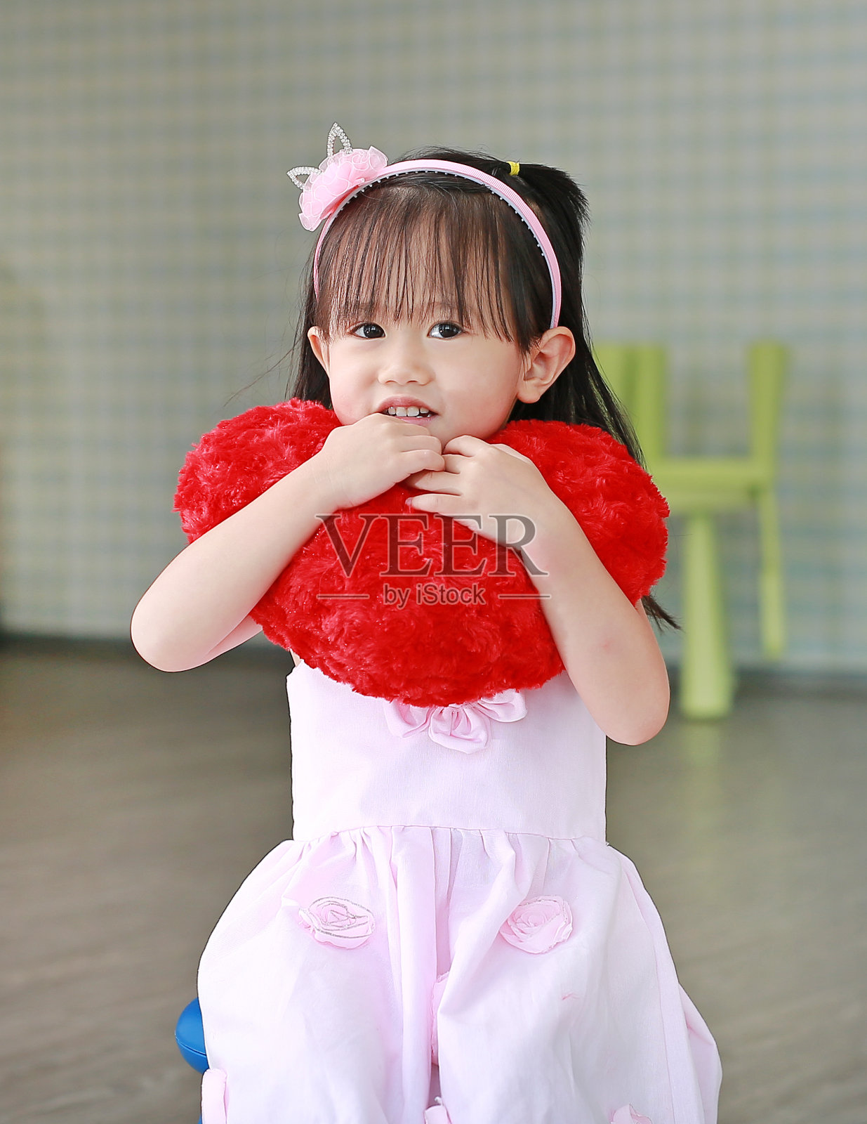 可爱的小女孩穿着粉红色的衣服坐在椅子上，红色的心枕头在游戏室。照片摄影图片