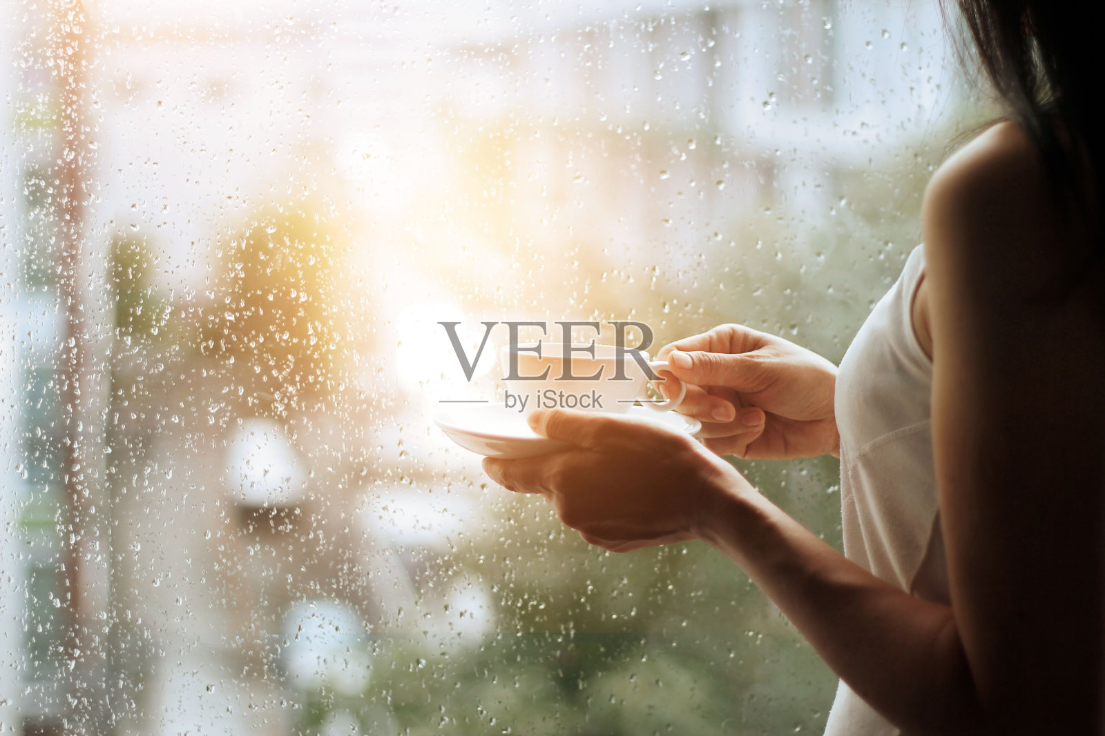 手里拿着咖啡的女人透过玻璃窗看着城市背景上的雨滴照片摄影图片
