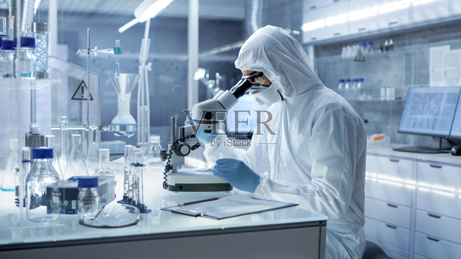 在一个安全的高水平研究实验室里，穿着工作服的科学家在显微镜下检查培养皿。照片摄影图片