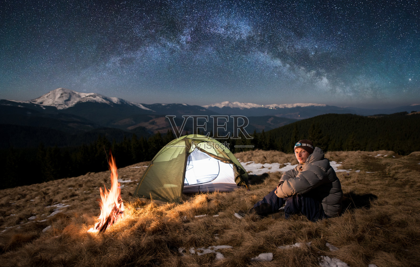男游客晚上在营地休息。坐在篝火和帐篷附近的人，看着镜头下的美丽的天空下，满是星星和银河。背景是白雪皑皑的群山照片摄影图片