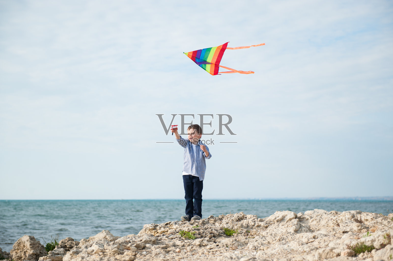 快乐的小男孩捧着风筝在天空中飞翔，背景是大海照片摄影图片
