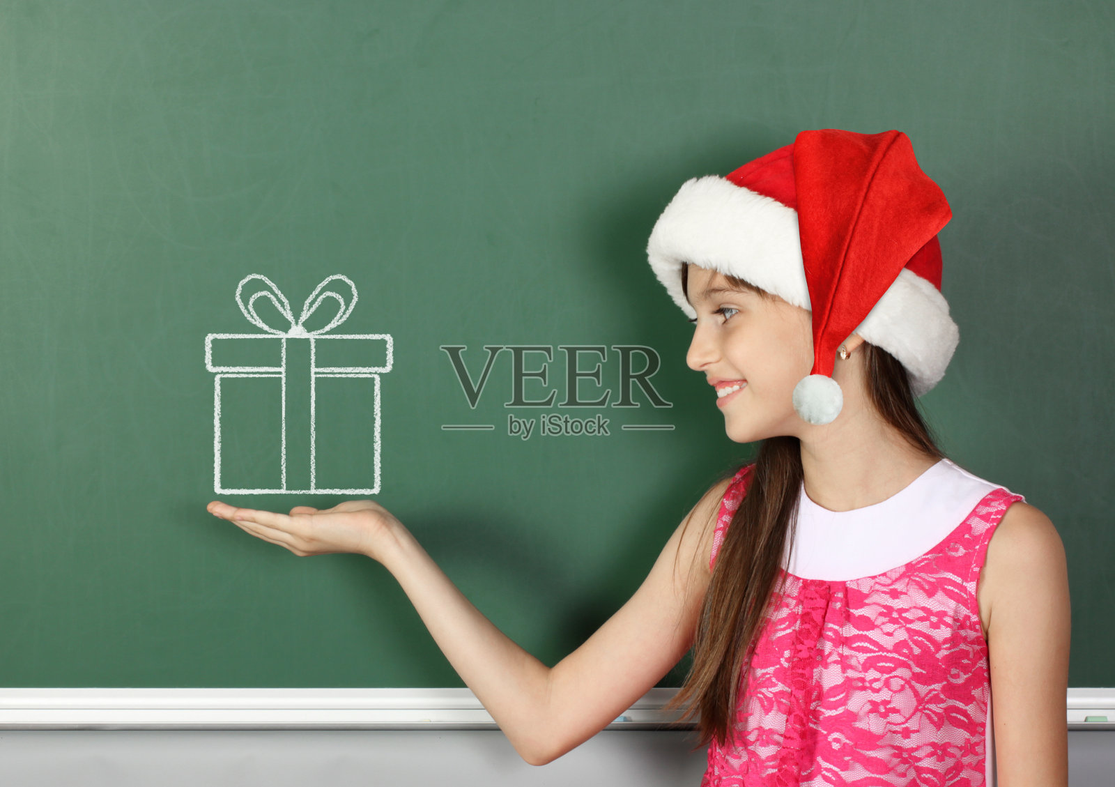 带着圣诞老人帽子的小女孩拿着画好的礼物盒靠近学校的黑板照片摄影图片