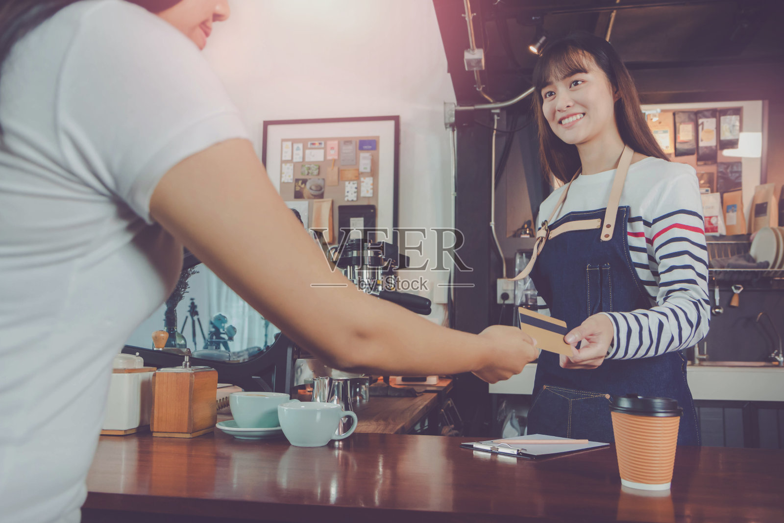 年轻美丽的亚洲女咖啡师穿着蓝色围裙，顾客在咖啡厅柜台用信用卡付款，面带微笑。咖啡店和小型咖啡店的概念。照片摄影图片