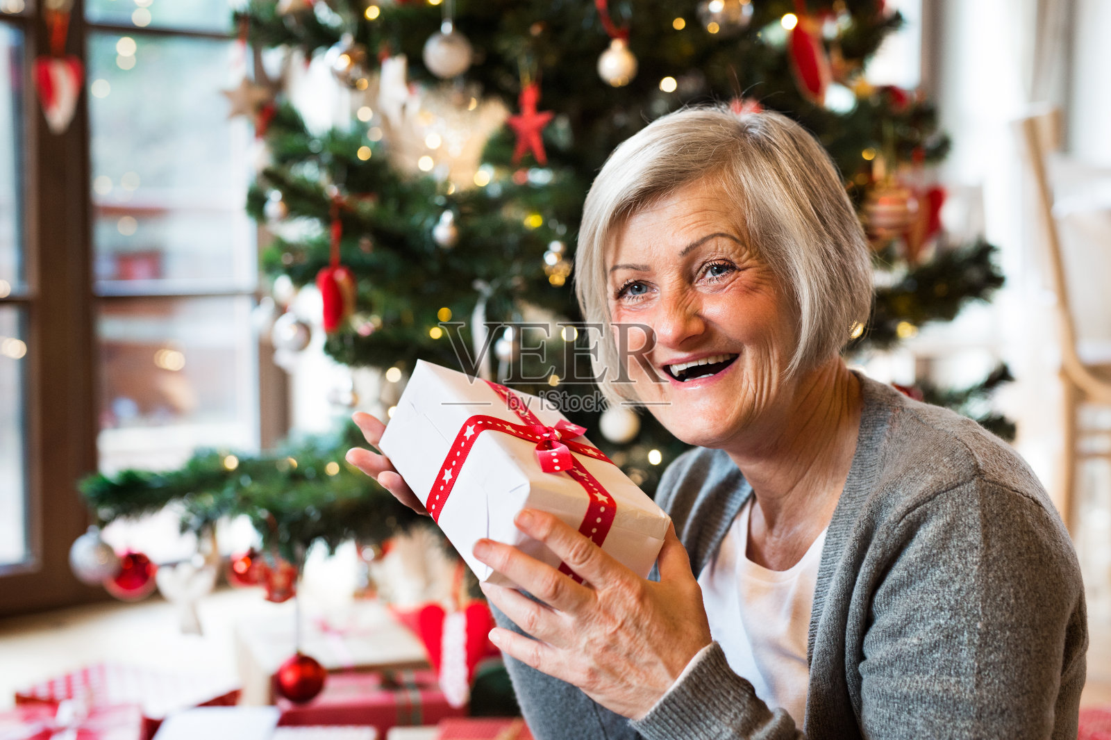 老妇人在圣诞树前拿着礼物。照片摄影图片