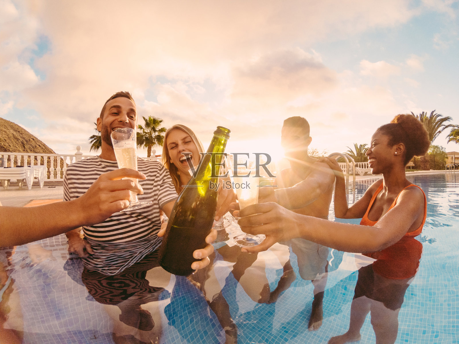 快乐的朋友欢呼与香槟在泳池派对在日落-富人有乐趣在独家热带度假-假日和友谊的概念-主要集中在左男人-太阳色调过滤器照片摄影图片