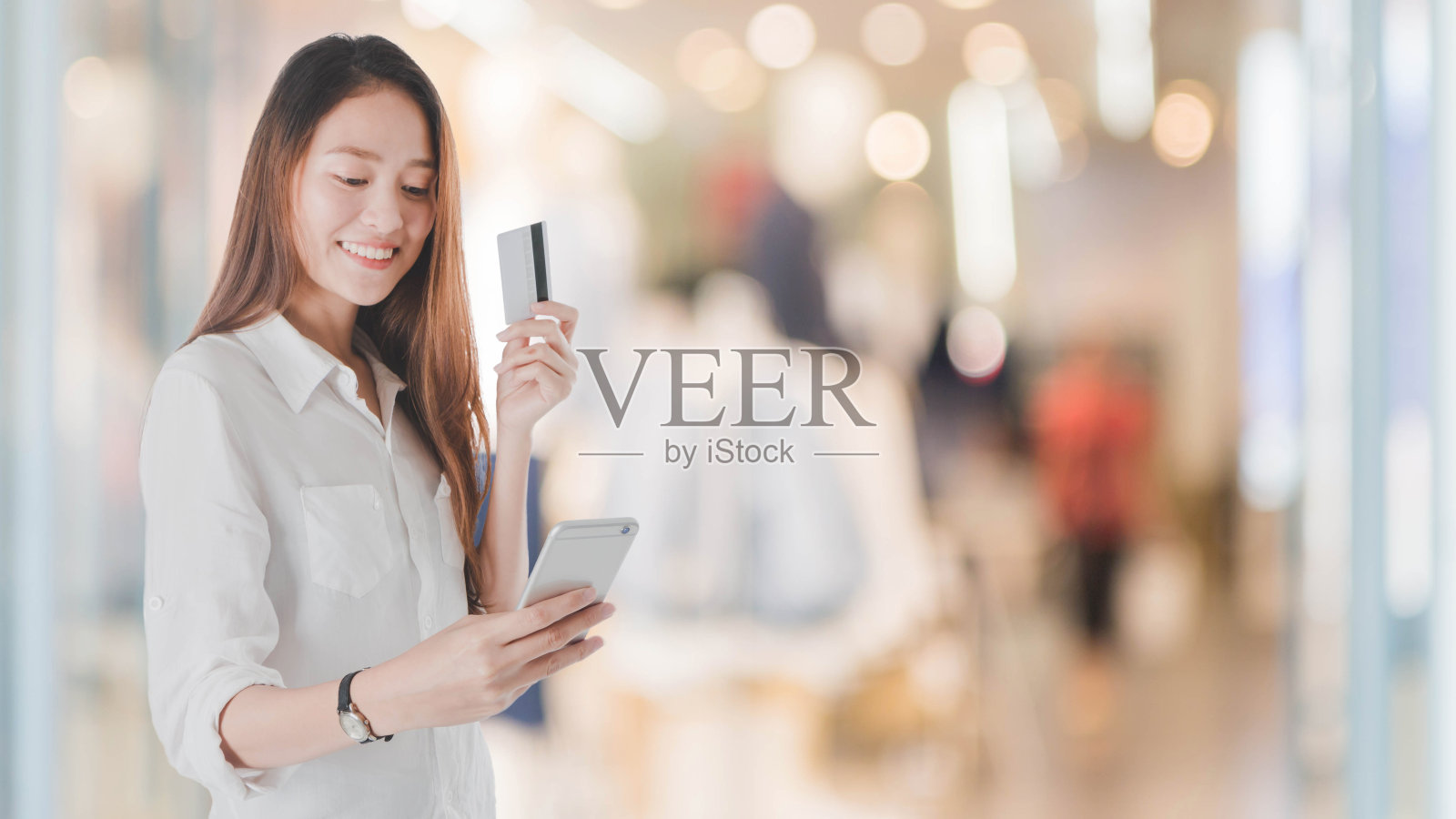 年轻美丽的亚洲女人用信用卡购物。在模糊的购物中心背景下拿着智能手机微笑。人们使用技术的概念。照片摄影图片