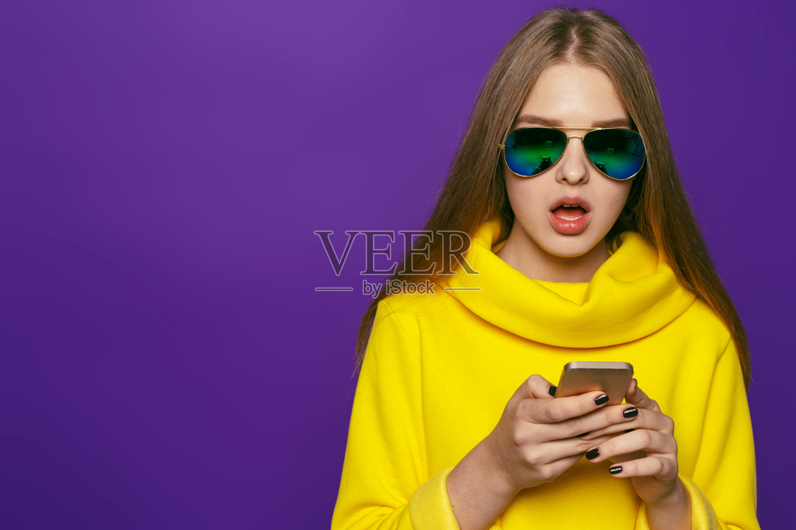 年轻的女孩手里拿着手机，很惊讶。在紫色的背景下穿着黄色的夹克照片摄影图片