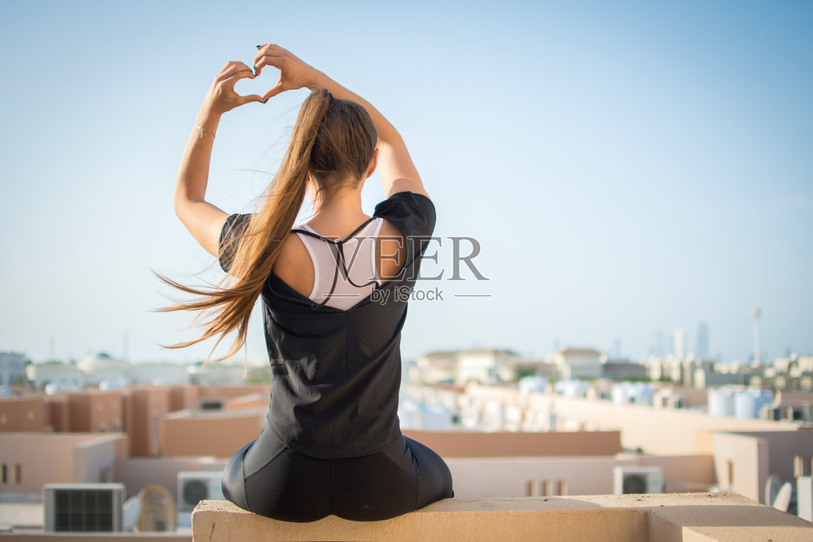 年轻女子坐在屋顶上，用手做心形的背影。照片摄影图片