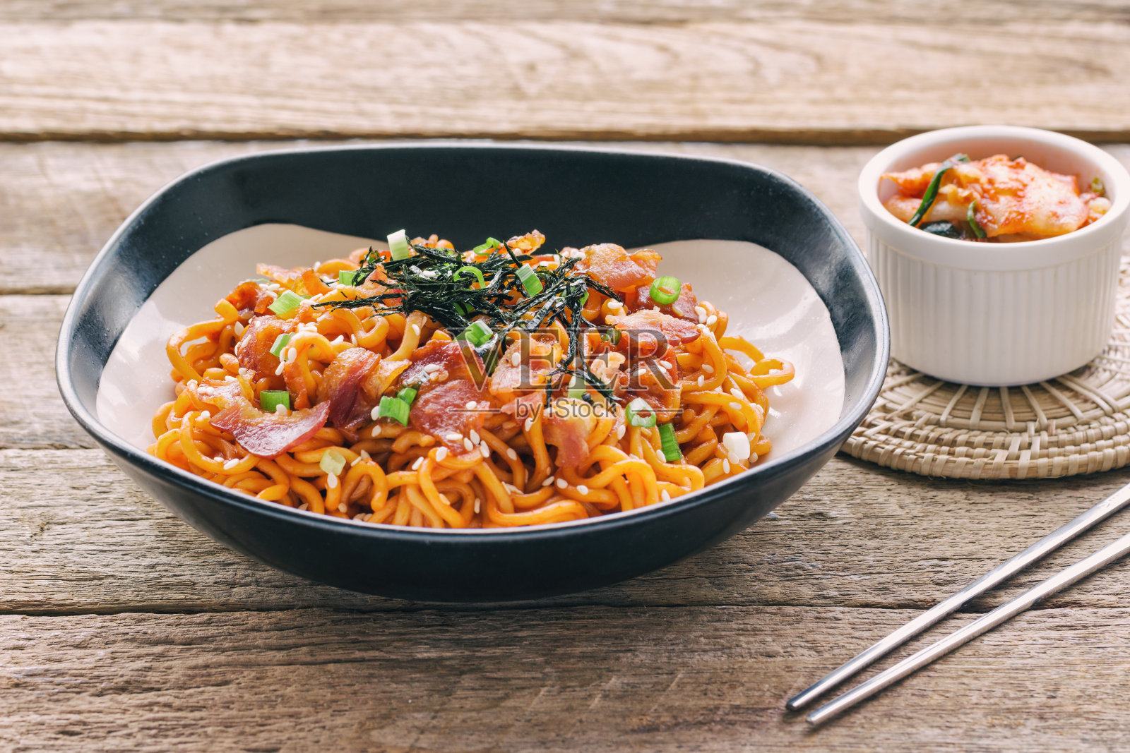 侧视图木桌上的黑白碗韩式辣味方便面，上面撒有培根、芝麻、海带、葱花和泡菜。自制美味亚洲食物的概念照片摄影图片