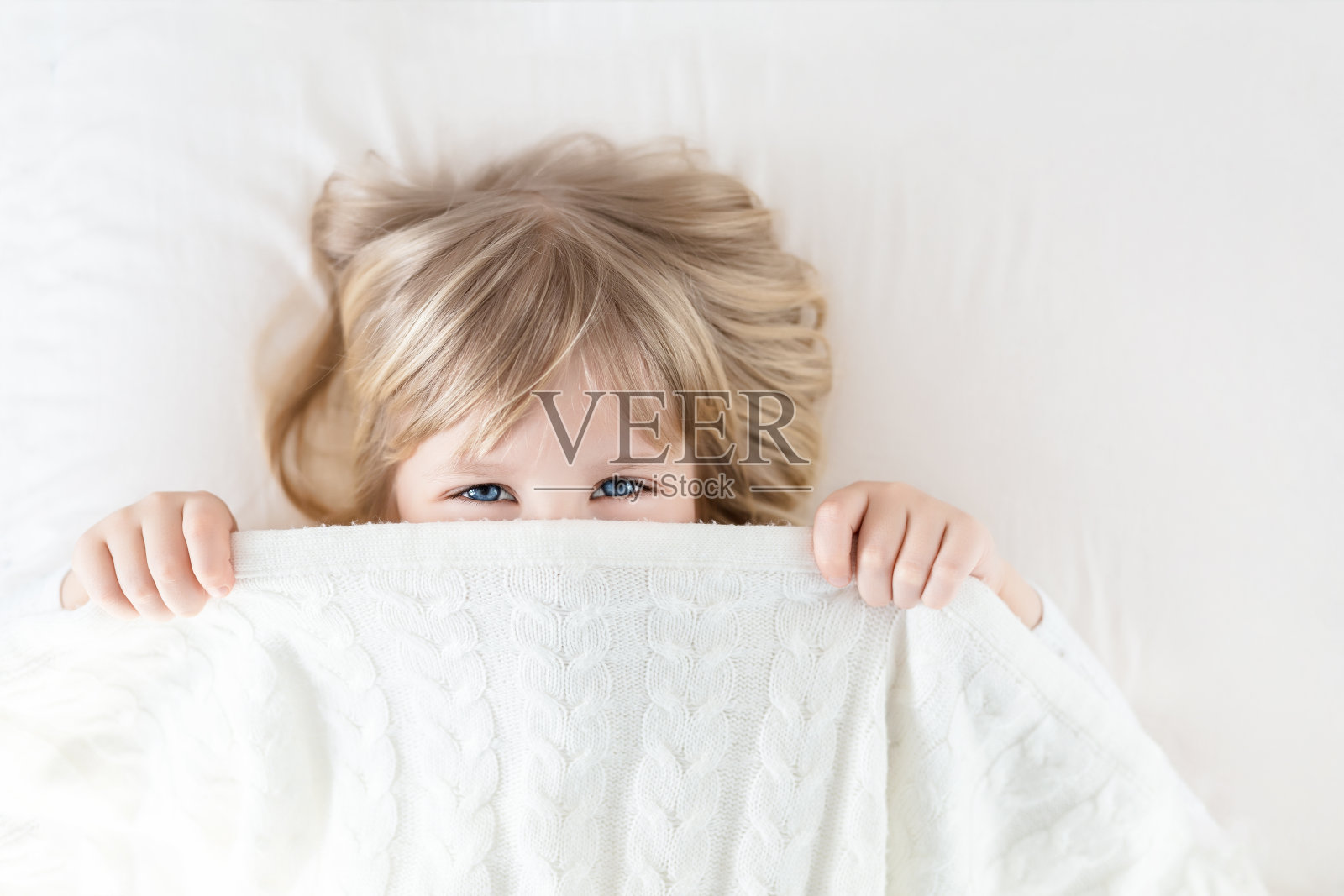小女孩从床上的毯子里往外偷看。可爱的孩子微笑着，藏在针织被子里。淘气的眼神。捉迷藏。孩子们玩得很开心。快乐的童年概念照片摄影图片