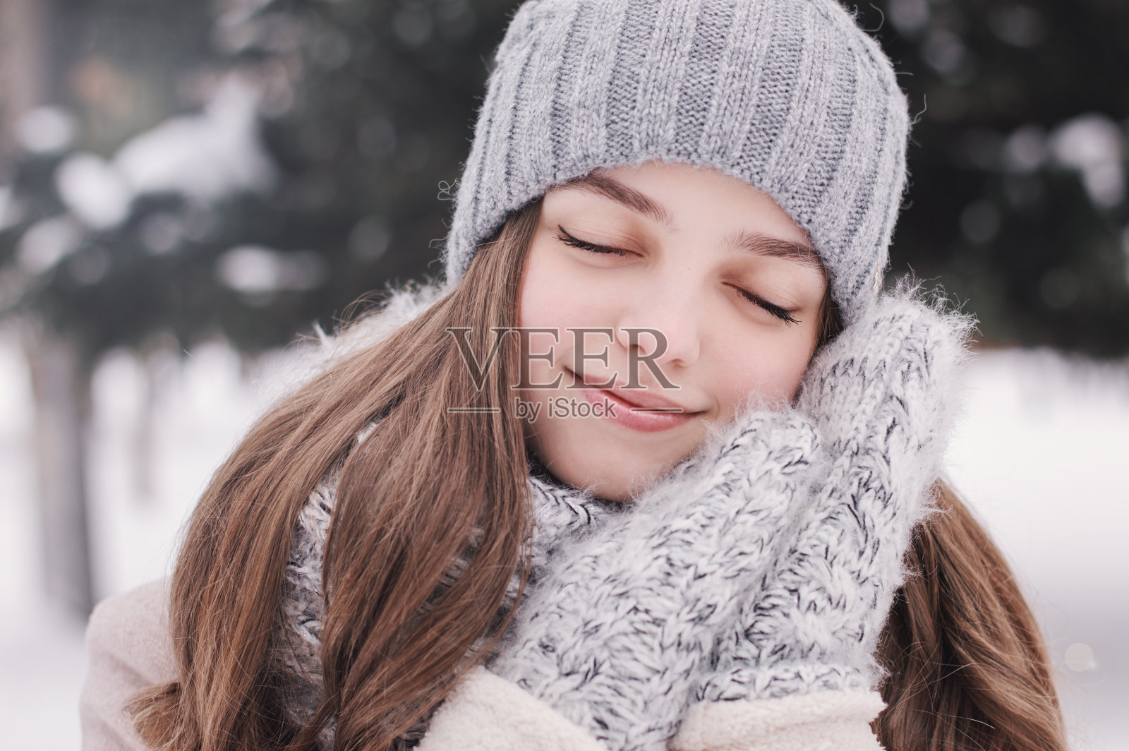 一个年轻女孩的横向肖像在灰色针织帽子，温暖的针织手套和米色外套。十几岁的女孩，闭着眼睛，双手放在脸旁边。积极的氛围。冬天的概念照片摄影图片