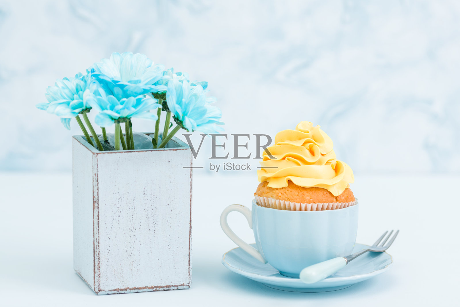 纸杯蛋糕与嫩黄奶油装饰和蓝色菊花花束复古破旧别致的花瓶在蓝色的粉彩背景。照片摄影图片