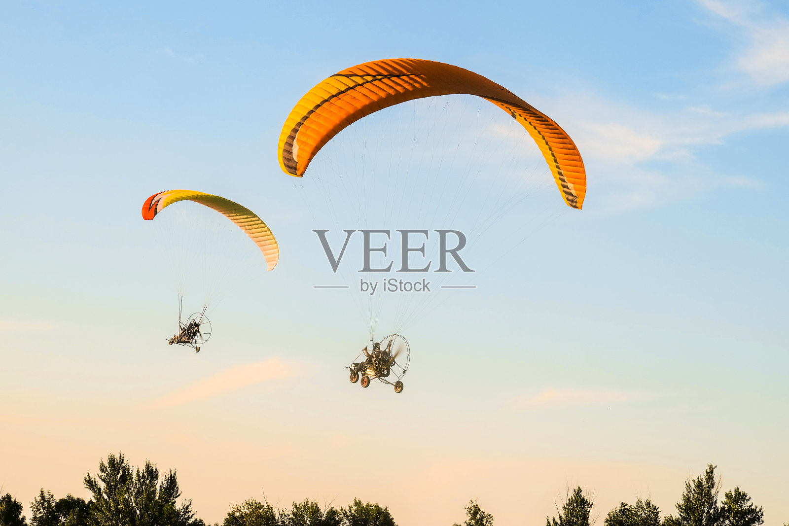 两个机动滑翔伞三轮车飞向天空。驾驶机动滑翔机在蓝天上飞过绿树。照片摄影图片